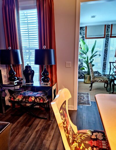 A modern living space with dark wood flooring, black lamps on a glass table, patterned bench and chair, and view into a sunlit room with plants and animal-print chairs.