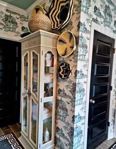 A white cabinet with glass doors stands against a patterned wallpapered wall, with woven baskets displayed on top and two black doors on either side.