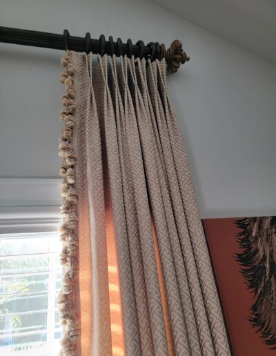 Beige patterned curtains with decorative trim hang from a dark rod near a window, partially illuminated by sunlight.