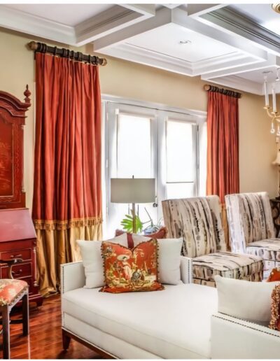 A traditional living room with patterned armchairs, a white sofa, orange drapes, a red cabinet, and decorative pillows in a bright, sunlit space.