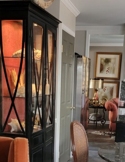 A living space with a black glass display cabinet holding shells, orange upholstered chairs, and framed artwork on cream walls in the background.