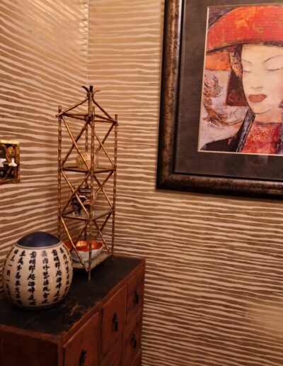 A small corner with textured wallpaper, a framed painting of a woman, a decorative vase with Asian script, a wooden chest, and a gold shelf with small ornaments.