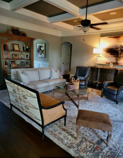 A living room with a beige sofa, two armchairs, a patterned bench, glass coffee table, bookshelf with decor, sideboard, artwork, lamp, ceiling fan, and area rug.