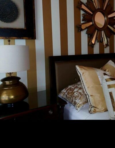 A bed with beige pillows, a dark headboard, a gold lamp on a nightstand, and a wall with gold and white vertical stripes and decorative mirrors.