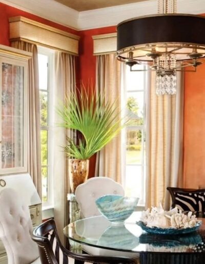 A dining room with orange walls, beige curtains, a glass table, upholstered chairs, decorative centerpiece items, a chandelier, and a glass-front cabinet.
